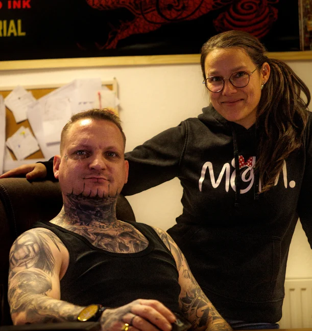 Tattoo-Studio-Inhaber und unterstützende Teammitglied im Bürobereich des Studios, vor einem roten Drachenmotiv, in einer warmen und einladenden Atmosphäre.
