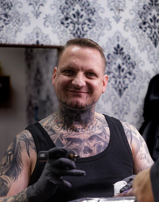 Ein Tattoo Artist mit Black-&-Grey-Realism-Fokus arbeitet lächelnd an einer Tattoo-Session im Studio, hält die Tattoo-Maschine bereit und trägt detailreiche Tattoos am Hals und Oberkörper.